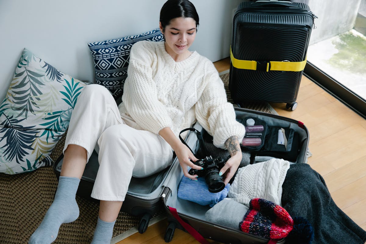 Can You Bring a Camera on a Plane: Guidelines for Travel Photographers 1 Young woman sitting on the floor packing a suitcase with clothes and a camera, preparing for a trip.