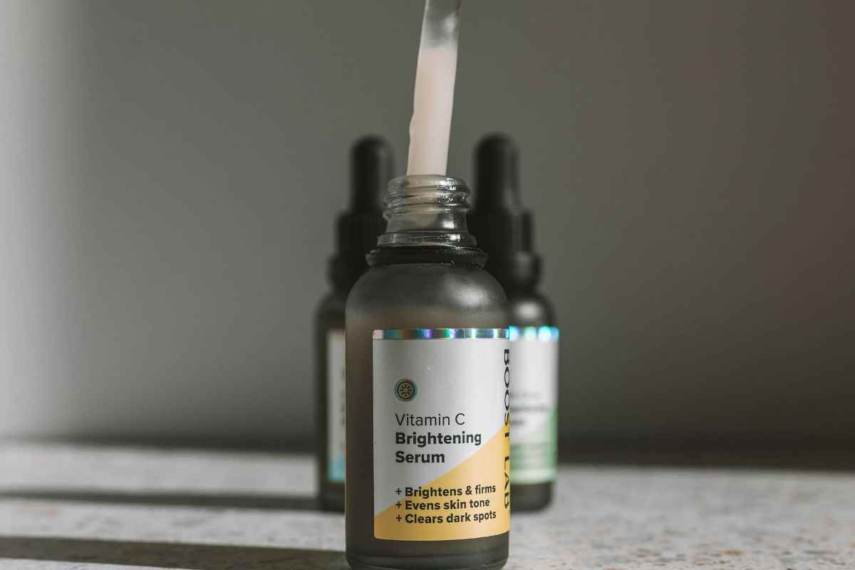 Close-up of a hand holding a dropper over a Vitamin C Brightening serum bottle on a textured surface.