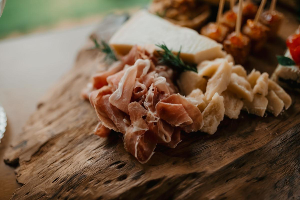 Folded prosciutto beside chunks of aged cheese and skewered bites on a rustic wooden charcuterie board