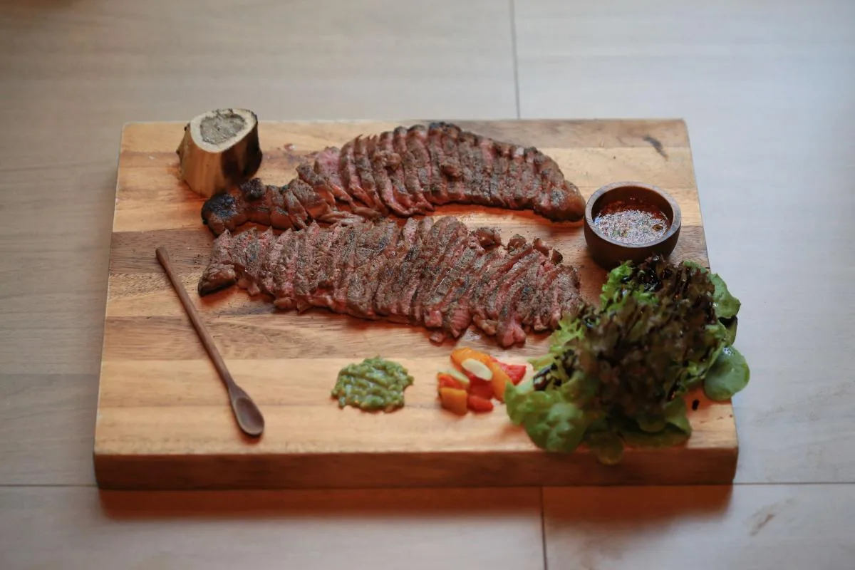 Sliced seared beef on a wooden cutting board with herbs, tomatoes and dipping sauce